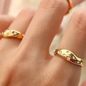 Puede incluir: Primer plano de anillos dorados con un dise&ntilde;o ondulado. Los anillos est&aacute;n adornados con peque&ntilde;as piedras preciosas transparentes. Los anillos est&aacute;n en los dedos de una persona, centr&aacute;ndose en la joyer&iacute;a.