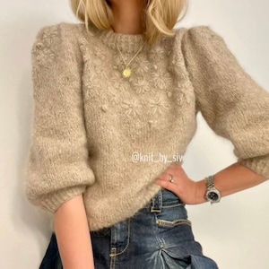 Könnte beinhalten: Ein beiger Strickpullover mit Blumenmuster und einer goldenen Halskette mit einem Sonnenanhänger. Der Pullover hat Puffärmel und eine lockere Passform. Der Text "@knit_by_siv" ist auf dem Pullover sichtbar.