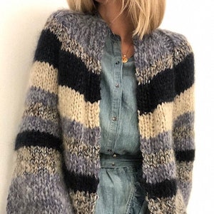 Puede incluir: Un cárdigan a rayas con una textura esponjosa en tonos beige, gris y negro. El cárdigan tiene un corte holgado y mangas largas.