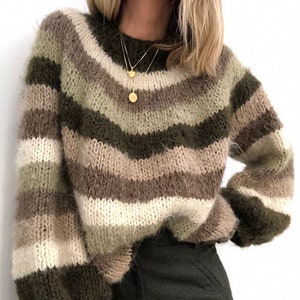 Puede incluir: Un suéter holgado con rayas horizontales en tonos verde oliva, marrón, beige y crema. El suéter tiene cuello redondo y mangas largas. La modelo lleva un collar dorado.