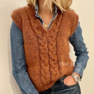 Pode incluir: Um colete de malha marrom sem mangas com gola em V e tranças. O colete é usado sobre uma camisa jeans azul e calças jeans. O texto "@knit_by_siv" é visível no canto inferior direito da imagem.
