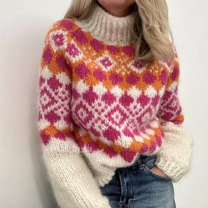 Könnte beinhalten: Ein weißer, rosa und orangefarbener Strickpullover mit geometrischem Muster. Der Pullover hat einen hohen Kragen und lange Ärmel.