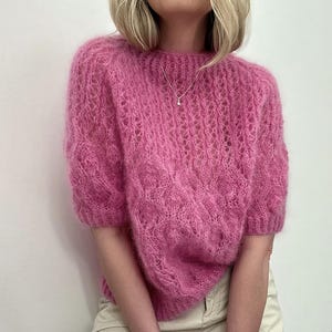 Puede incluir: Un suéter rosa brillante de manga corta con un diseño de punto abierto texturizado. El suéter tiene cuello redondo y un aspecto suave y difuso. La prenda está hecha de un material ligero, perfecto para combinar.