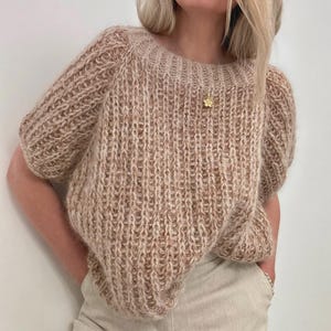 Puede incluir: Un suéter beige de manga corta con un diseño de punto acanalado. El suéter tiene un cuello redondo y mangas ligeramente abullonadas. Un colgante dorado cuelga de una delicada cadena. El suéter está hecho de un hilo suave y texturizado.