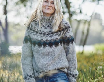May include: A light gray knit sweater featuring a high neck and a dark geometric pattern across the chest and cuffs. The sweater is paired with blue denim jeans. The knit texture is visible throughout the garment, showcasing a thick yarn construction.