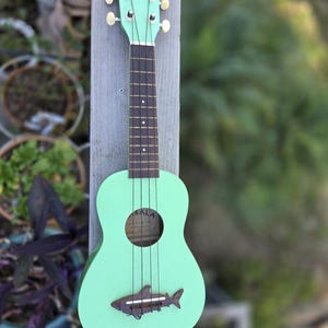 May include: A mint green ukulele with the word "MAKALA" on the headstock. The ukulele has a shark-shaped soundhole decoration and black strings. The instrument is displayed outdoors, leaning against a wooden post.