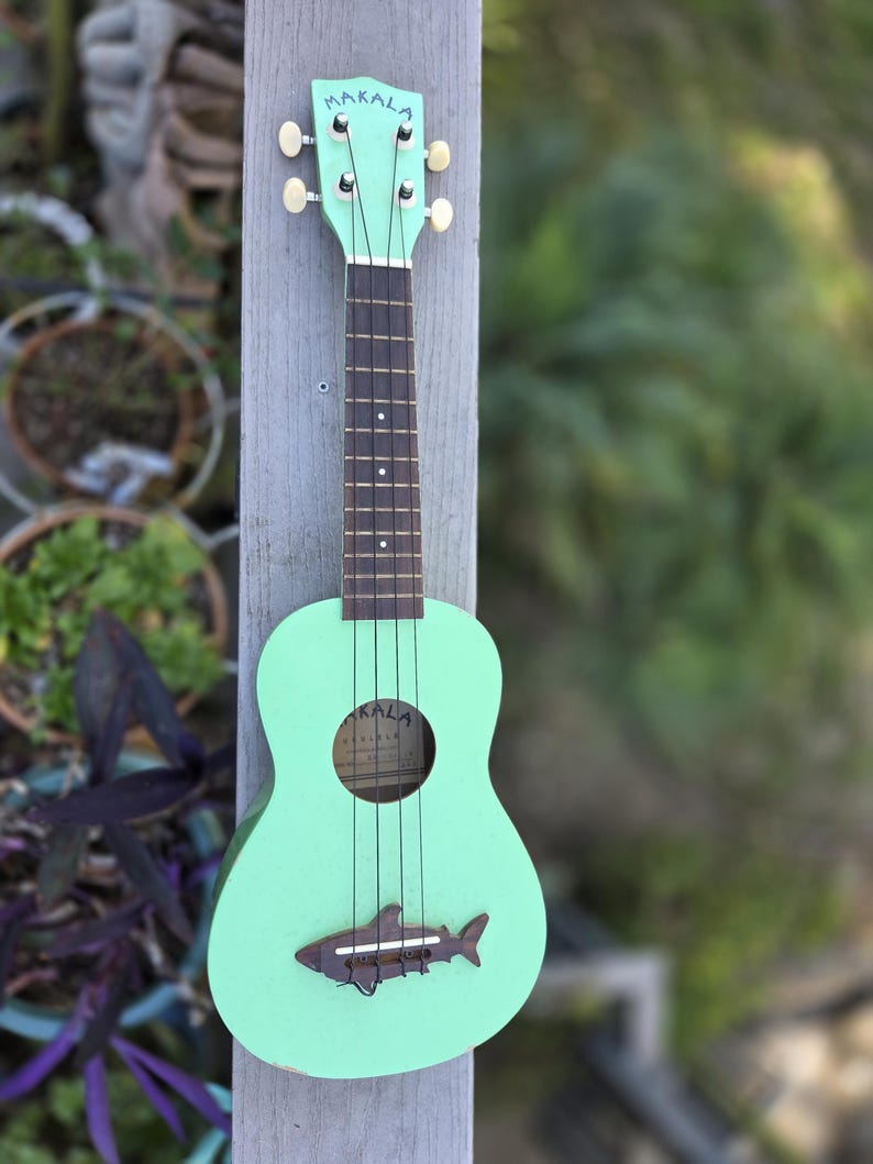 May include: A mint green ukulele with the word "MAKALA" on the headstock. The ukulele has a shark-shaped soundhole decoration and black strings. The instrument is displayed outdoors, leaning against a wooden post.