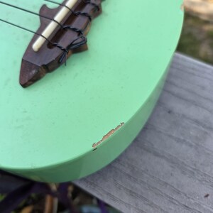 May include: A mint green ukulele with a shark-shaped bridge. The bridge is made of dark wood and holds four black strings. The ukulele's body shows some wear. The instrument is resting on a wooden surface.
