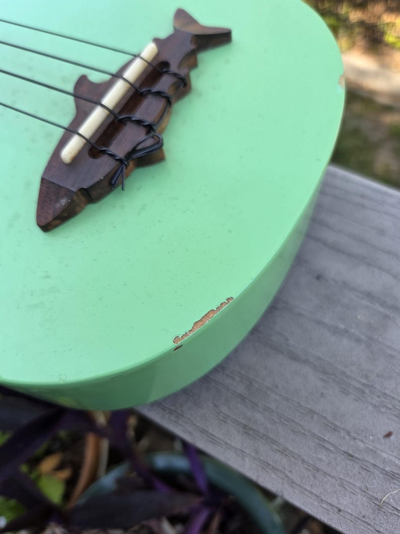 May include: A mint green ukulele with a shark-shaped bridge. The bridge is made of dark wood and holds four black strings. The ukulele's body shows some wear. The instrument is resting on a wooden surface.