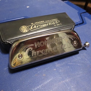 May include: A silver Hohner Chrometta 8 harmonica with a black case. The harmonica has the name "Hohner Chrometta 8" engraved on it. The case is black with gold lettering that reads "M. Hohner Made in Germany Chrometta 8 Nr. 250 C".