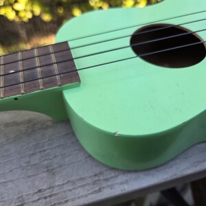 May include: A light green ukulele with black strings and a dark brown fretboard. The instrument has a round sound hole and is resting on a wooden surface. The ukulele's body shows signs of wear.