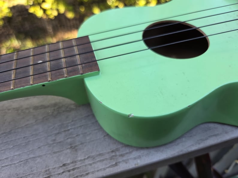 May include: A light green ukulele with black strings and a dark brown fretboard. The instrument has a round sound hole and is resting on a wooden surface. The ukulele's body shows signs of wear.