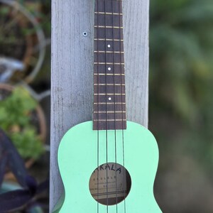May include: A mint green ukulele with the word "MAKALA" on the headstock. The ukulele has a shark-shaped bridge and the word "UKULELE" inside the sound hole. The instrument is mounted on a wooden post.