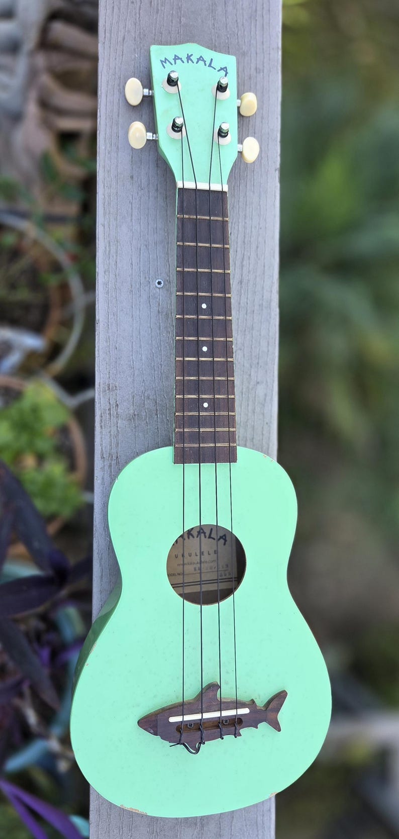 May include: A mint green ukulele with the word "MAKALA" on the headstock. The ukulele has a shark-shaped bridge and the word "UKULELE" inside the sound hole. The instrument is mounted on a wooden post.