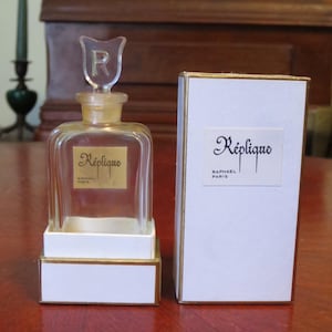 May include: A vintage perfume bottle and box set. The clear glass bottle has a unique stopper with an "R" design. The box and the bottle's label both feature the name "Réplique" in elegant script, with "Raphael Paris" below.