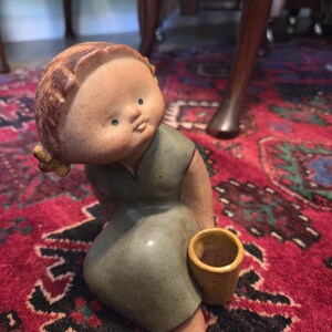 May include: A ceramic figurine of a seated child wearing a green dress and holding a small woven basket. The figurine has a detailed face with brown hair and is set against a red patterned rug.