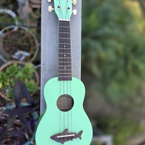 May include: A mint green ukulele with the word "MAKALA" on the headstock. The ukulele has a shark-shaped bridge and four strings. The instrument is leaning against a wooden post, with greenery in the background.