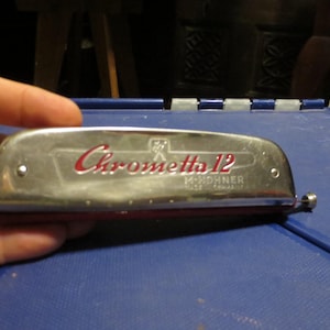 May include: A silver Chrometta 12 harmonica with red lettering and accents. The harmonica is made by M. Hohner, Made in Germany. The instrument is held against a blue surface.