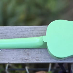May include: A mint green ukulele, seen from the back, on a wooden surface. The body and neck are a solid, pastel green. Silver tuning pegs with cream-coloured knobs are visible. The ukulele is a stringed instrument.