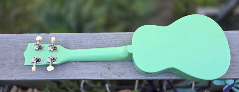 May include: A mint green ukulele, seen from the back, on a wooden surface. The body and neck are a solid, pastel green. Silver tuning pegs with cream-coloured knobs are visible. The ukulele is a stringed instrument.