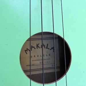 May include: A close-up shot of a mint green ukulele with black strings. The sound hole displays the brand name "MAKALA UKULELE" and website address. The ukulele's body shows signs of wear, adding to its vintage charm.