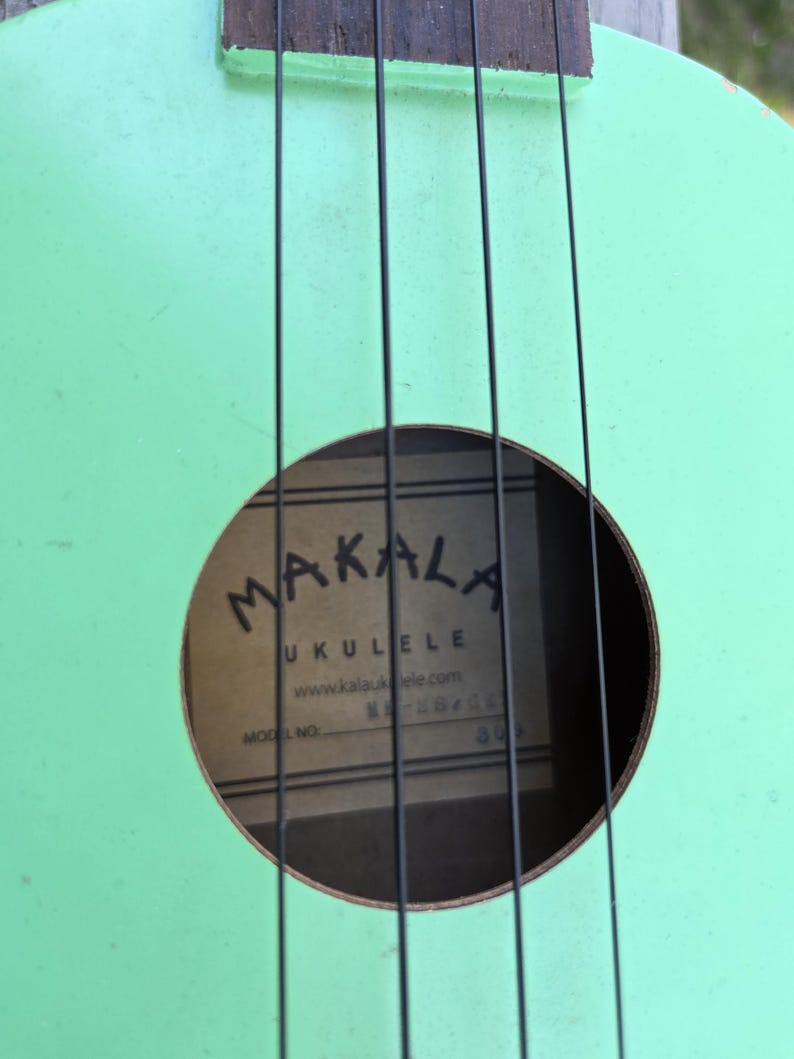 May include: A close-up shot of a mint green ukulele with black strings. The sound hole displays the brand name "MAKALA UKULELE" and website address. The ukulele's body shows signs of wear, adding to its vintage charm.