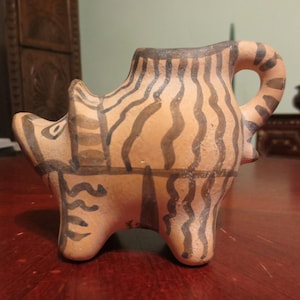 May include: A tan ceramic figurine of an animal with dark brown stripes. The figurine has a handle and is standing on four legs. The stripes are wavy and cover the body of the animal. The figurine is likely a decorative item.