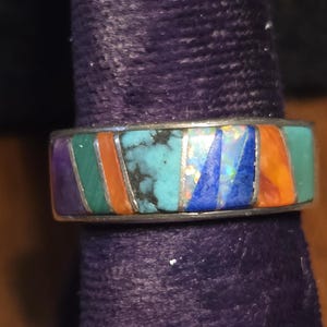 May include: A silver ring with a colorful inlaid design. The ring features rectangular sections of turquoise, opal, lapis lazuli, orange, green, and purple stones. The ring is set against a dark purple background.