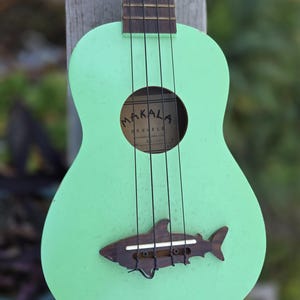 May include: A mint green ukulele with a shark-shaped bridge. The ukulele features black strings and a sound hole with the brand name "Makala". The instrument is made of wood and has a distressed finish, suggesting a vintage or handmade aesthetic.