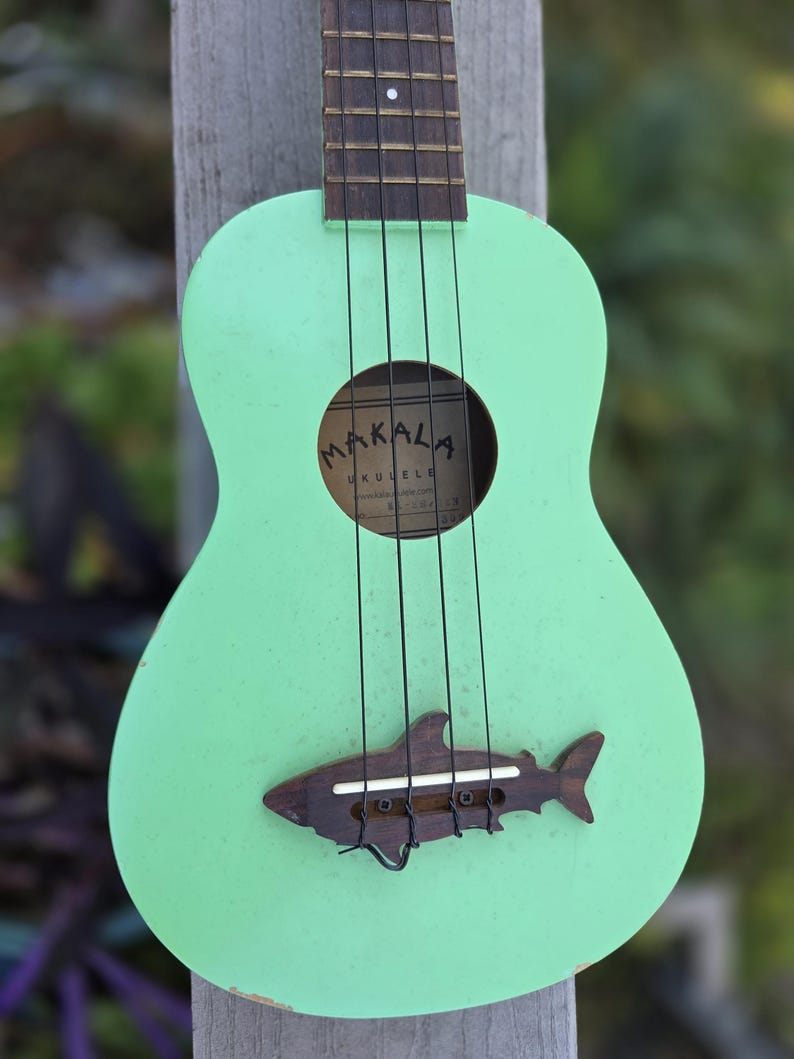 May include: A mint green ukulele with a shark-shaped bridge. The ukulele features black strings and a sound hole with the brand name "Makala". The instrument is made of wood and has a distressed finish, suggesting a vintage or handmade aesthetic.