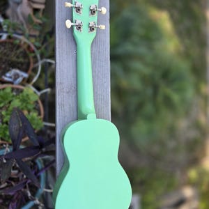 May include: A mint green ukulele, seen from the rear, is mounted on a weathered wooden post. The instrument's body and neck are a solid pastel green, with silver tuning pegs and cream-coloured knobs. The background is a soft blur of green foliage.