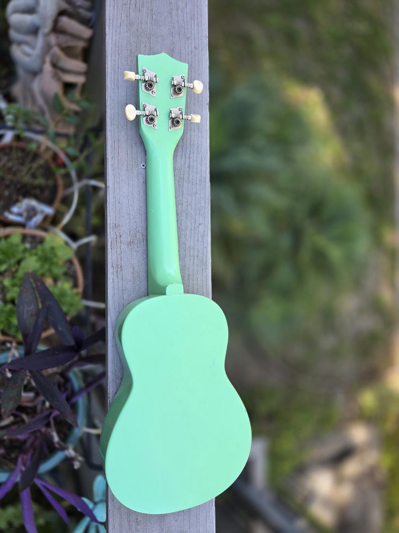 May include: A mint green ukulele, seen from the rear, is mounted on a weathered wooden post. The instrument's body and neck are a solid pastel green, with silver tuning pegs and cream-coloured knobs. The background is a soft blur of green foliage.