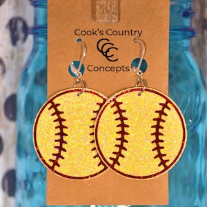 May include: A pair of softball earrings with glitter and red stitching. The earrings are on a brown card with the text "Cook's Country Concepts".