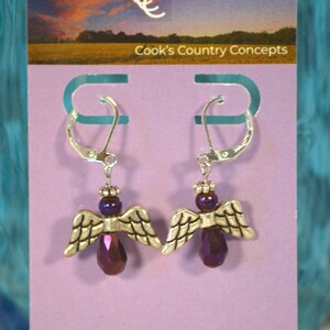May include: Purple crystal angel wing earrings with silver hooks. The earrings are displayed on a card with the text "Cook's Country Concepts" and website information.