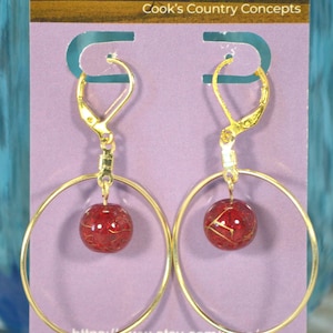 May include: A pair of gold-colored hoop earrings with a red, spherical bead. The beads have a gold-colored, web-like design inside. The earrings are displayed on a purple card with the text "Cook's Country Concepts".