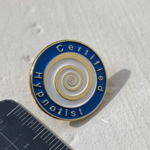 Peut inclure: Une épingle en émail bleu et blanc avec les mots "Hypnotiseur certifié" et un motif en spirale doré.
