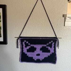 May include: A black and purple crochet wall hanging featuring a cat skull design. The handmade piece is suspended from a wooden dowel with a black and gray cord. The cat skull is in shades of purple.
