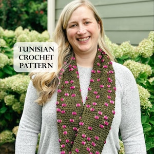 May include: A woman wearing a green Tunisian crochet infinity scarf with pink accents. The scarf is made of a textured yarn and features a repeating pattern of pink bobbles. The text "TUNISIAN CROCHET PATTERN" is in a white box in the image.