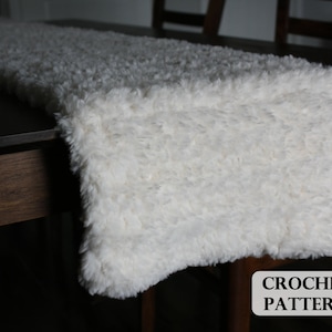 May include: A white crocheted table runner with a fluffy texture. The runner is laying on a dark wood table. CROCHET PATTERN is written in black text on the table.