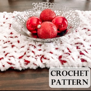 May include: A white and red crochet table runner with a silver bowl containing four red Christmas ornaments. The bowl has a decorative pattern with numbers. The text "CROCHET PATTERN" is in a white box.