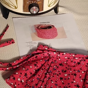 Puede incluir: Una cubierta para olla de cocción lenta de tela roja y blanca con un patrón de estrellas. La cubierta tiene un cierre de cordón y se muestra en la parte superior de una olla de cocción lenta.