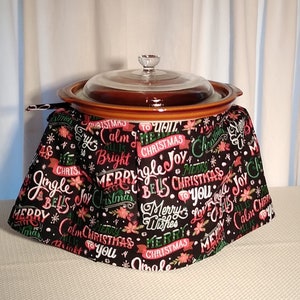 Puede incluir: Una olla de cocción lenta marrón con una tapa de vidrio, cubierta con una tela negra con un estampado navideño. El estampado presenta texto rojo y verde y copos de nieve. El texto incluye frases como "Jingle Bells", "Feliz Navidad" y "Calm All Is Bright".