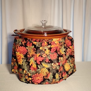 May include: A brown slow cooker with a glass lid and a floral patterned fabric cover. The cover features a black background with red, yellow, and green flowers and leaves.
