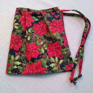 Puede incluir: Una bolsa con cordón con un diseño de flor de pascua navideña. La bolsa muestra grandes flores de pascua rojas, agujas de pino verdes y piñas sobre un fondo oscuro. El cordón es rojo y verde.