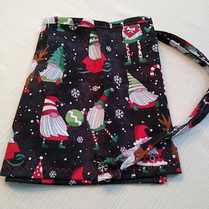 Puede incluir: Un delantal de tela negro con un patrón de gnomo navideño. El delantal tiene un borde rojo y una cuerda para atar. Los gnomos llevan gorros rojos, barbas blancas y pantalones verdes. El fondo es negro con copos de nieve blancos.