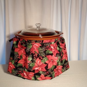 May include: A brown slow cooker with a glass lid, covered in a black and red fabric with a poinsettia pattern. The fabric has a gathered skirt around the base of the cooker.