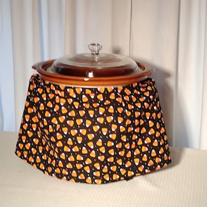 Puede incluir: Una olla de cocción lenta marrón con una tapa de vidrio, cubierta con una tela negra con un patrón de maíz dulce naranja y blanco.