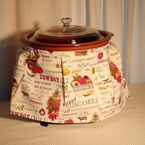 Puede incluir: Una olla de cocción lenta marrón con una tapa de vidrio, cubierta con una tela blanca con un estampado colorido de varios ingredientes y especias. La tela tiene texto que dice "Cowboy Chili" y "Kickin' Guacamole".
