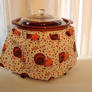 Puede incluir: Una olla de cocción lenta marrón con una tapa de vidrio, cubierta con una tela blanca con un tema de Acción de Gracias. La tela tiene un patrón de pavos, calabazas, hojas y peras.