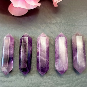 May include: Five purple amethyst crystal points, each with a smooth, polished finish. The points are arranged in a row on a gray textured surface.